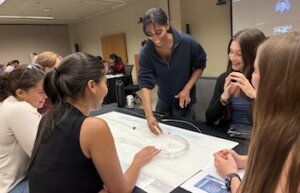 A group of young interns lean in around a table as they watch a demonstration of the Seldinger technique on a 3D simulation model.
