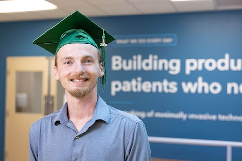 Cook Medical employee Duncan Gage stands in front of inspirational signage wearing his HSE graduation cap